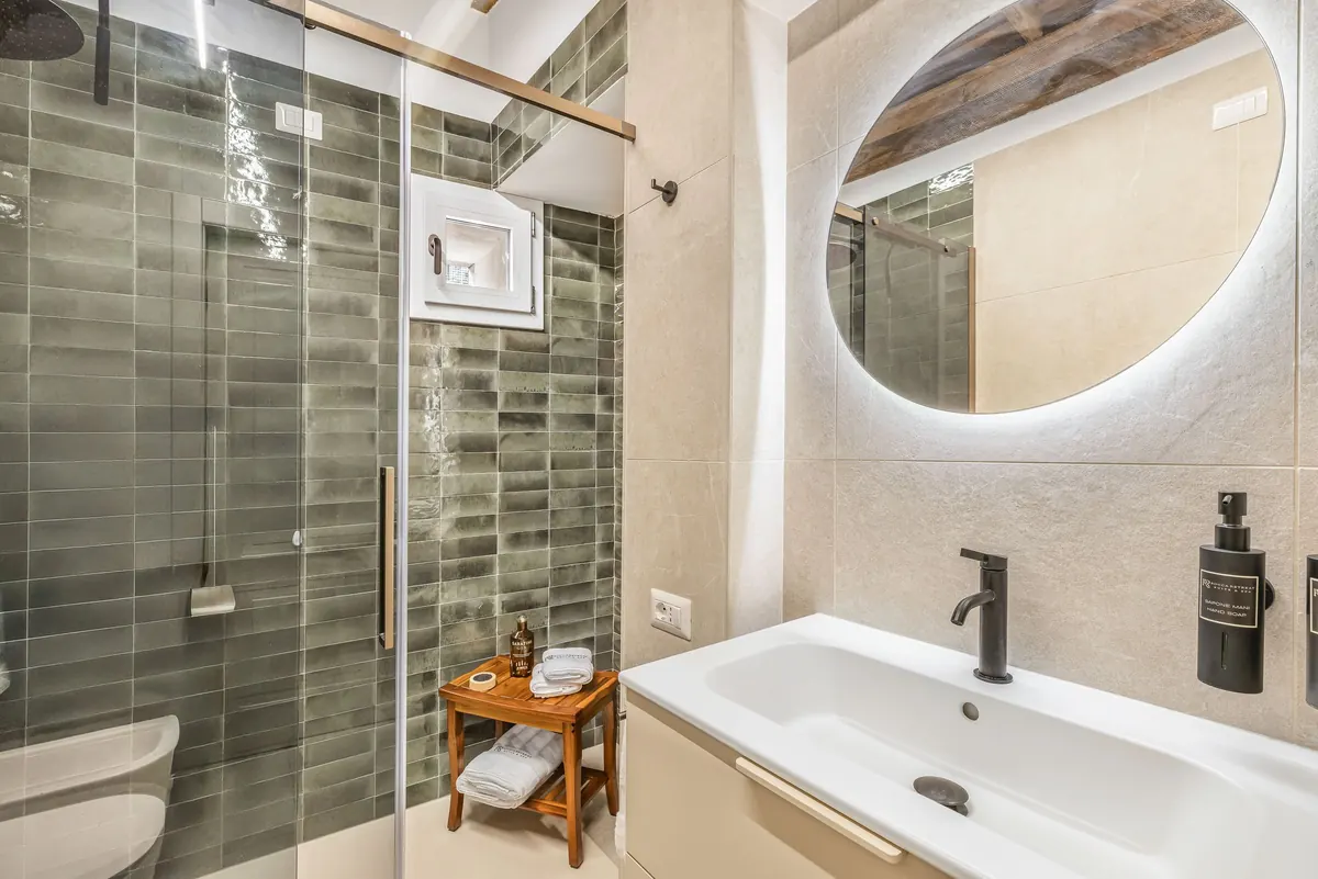 Bagno con doccia in vetro, piastrelle verde salvia e specchio tondo retroilluminato